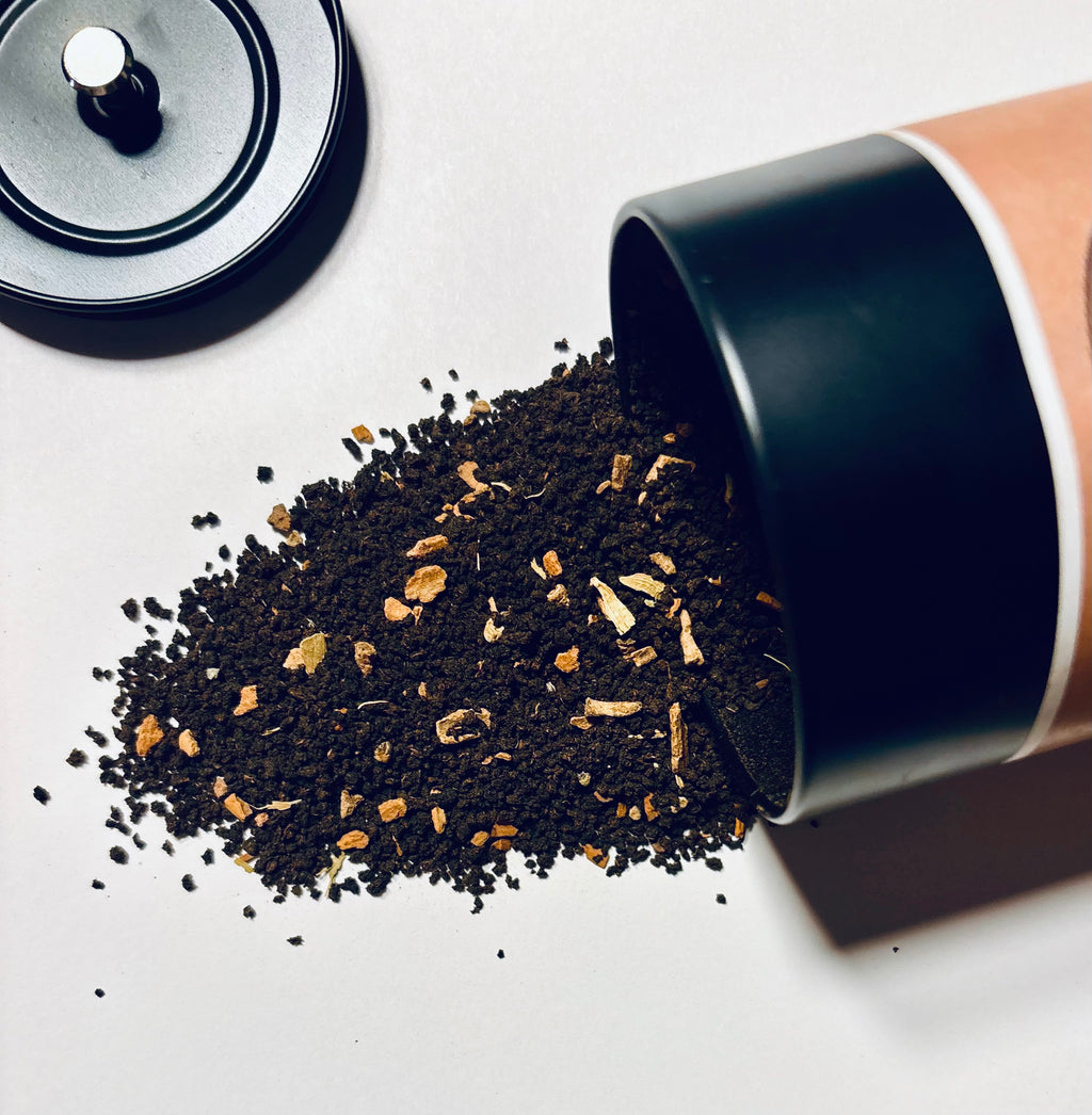 A close-up of loose dark coffee grounds or tea spilling from a black-and-tan cylindrical container onto a light surface, with the container lid visible in the upper left.